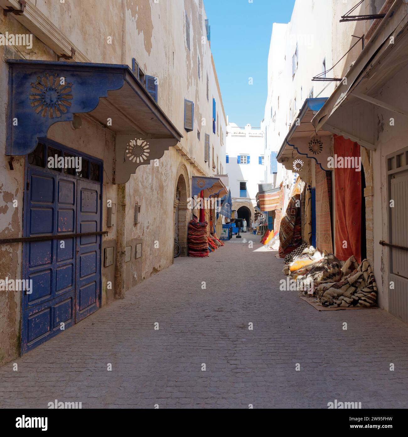 Rue étroite dans la médina historique avec porte bleue et piles de tapis prêt à être vendu à Essaouira, Maroc. 23 décembre 2023 Banque D'Images