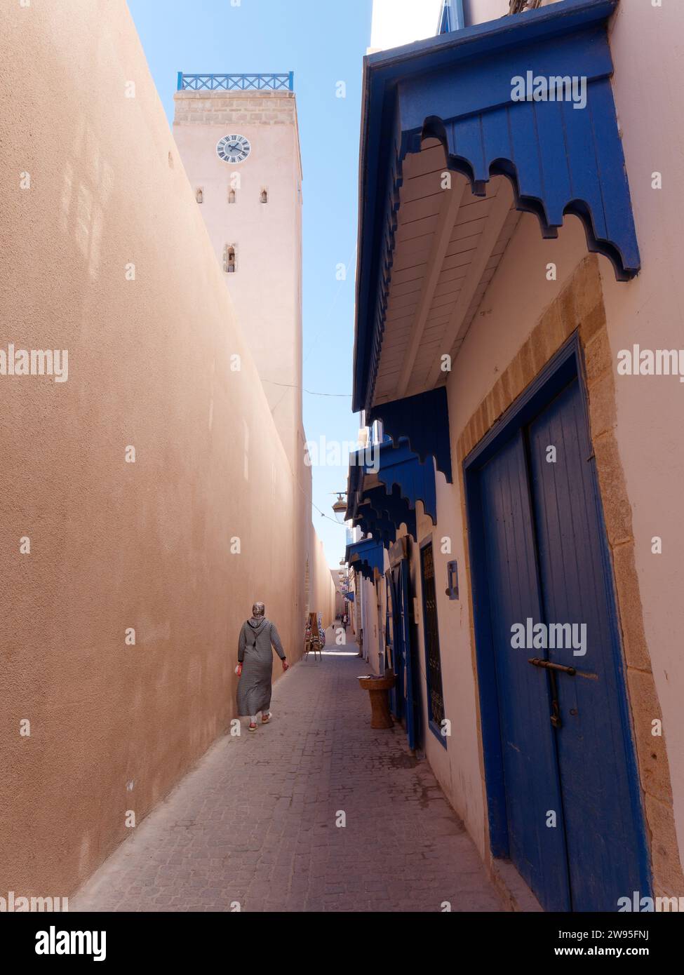 Rue étroite dans la médina historique avec porte bleue et une tour de l'horloge comme une femme musulmane passe à Essaouira, Maroc. 23 décembre 2023 Banque D'Images