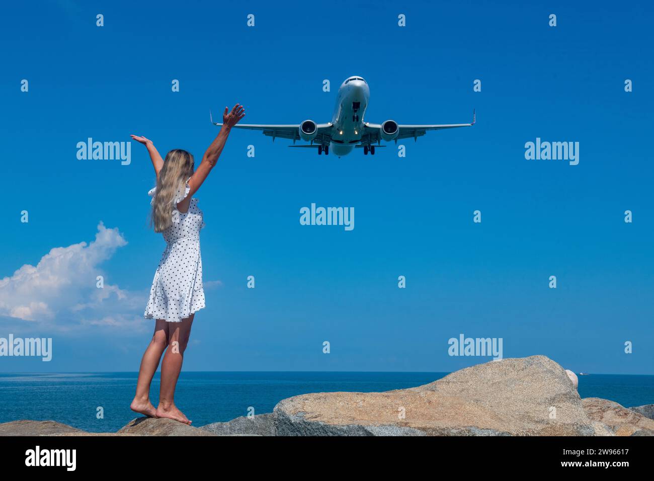 Aerial Rendezvous : fille en robe blanche sur pierres, avion au-dessus de la mer Bleue Banque D'Images