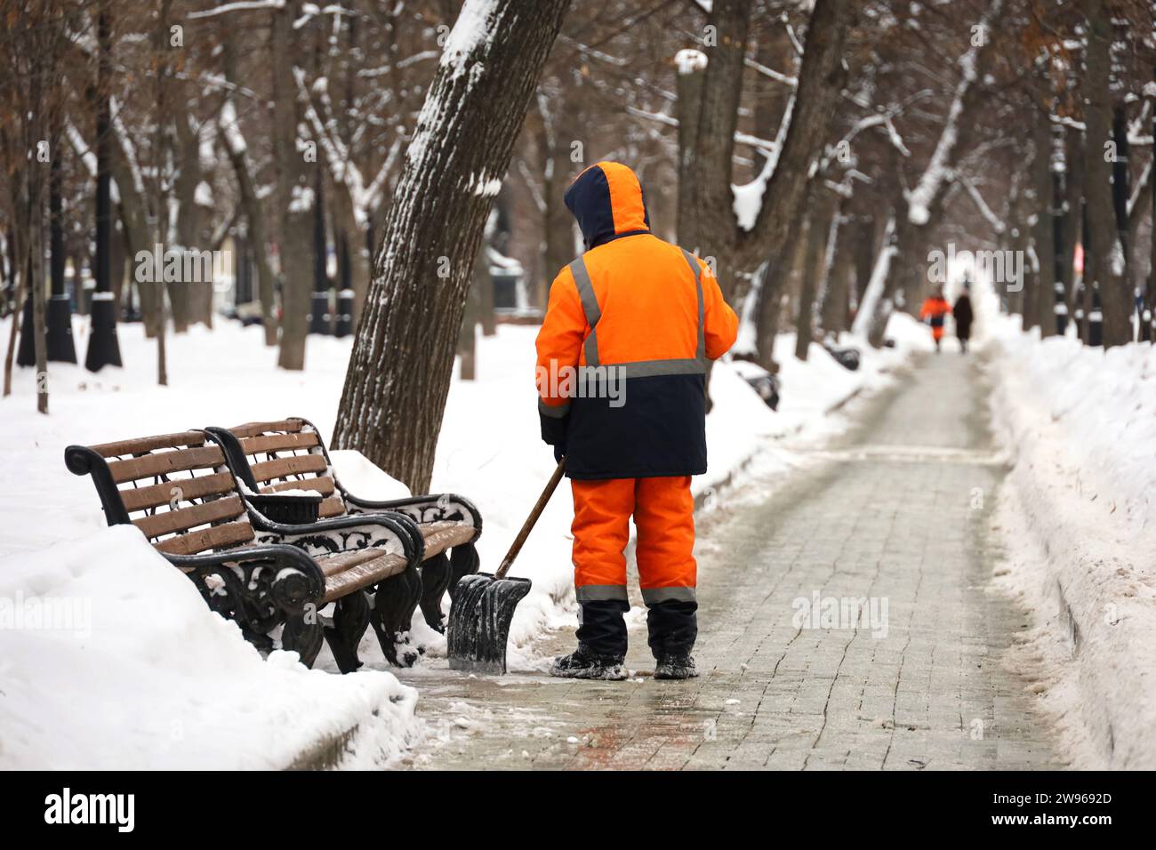 Travailleur des services communautaires en uniforme avec une pelle déneige sur un trottoir. Homme pendant le déneigement dans la ville d'hiver, nettoyage des rues Banque D'Images
