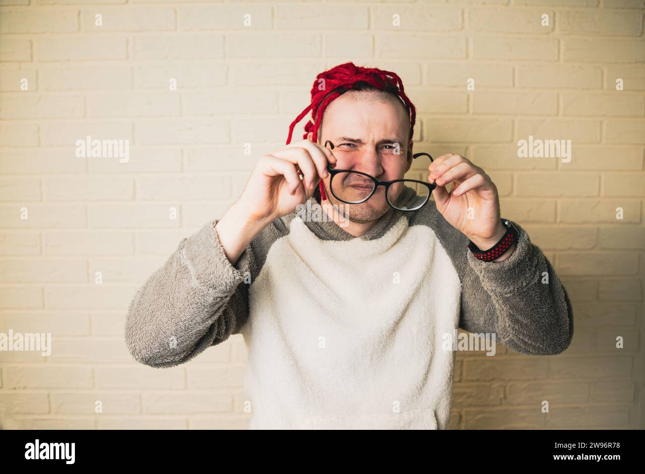 Un bel homme adulte avec des lunettes et des dreadlocks rouges plisse et regarde la caméra contre un mur de briques blanches. Banque D'Images
