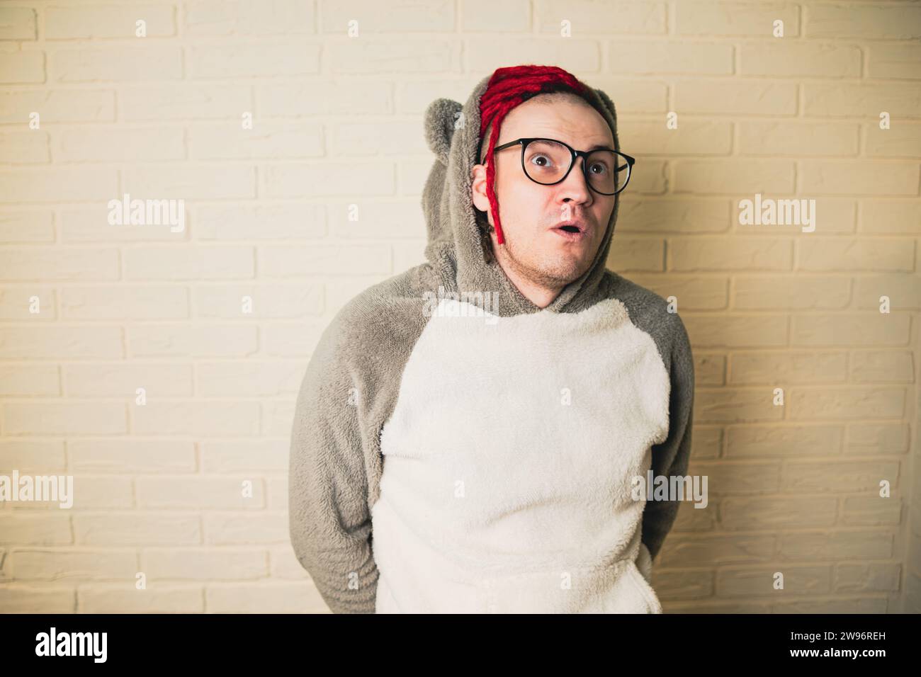 Un bel homme adulte avec des lunettes et des dreadlocks rouges regarde avec intérêt la pièce autour de lui sur le fond d'un mur de briques blanches. Banque D'Images