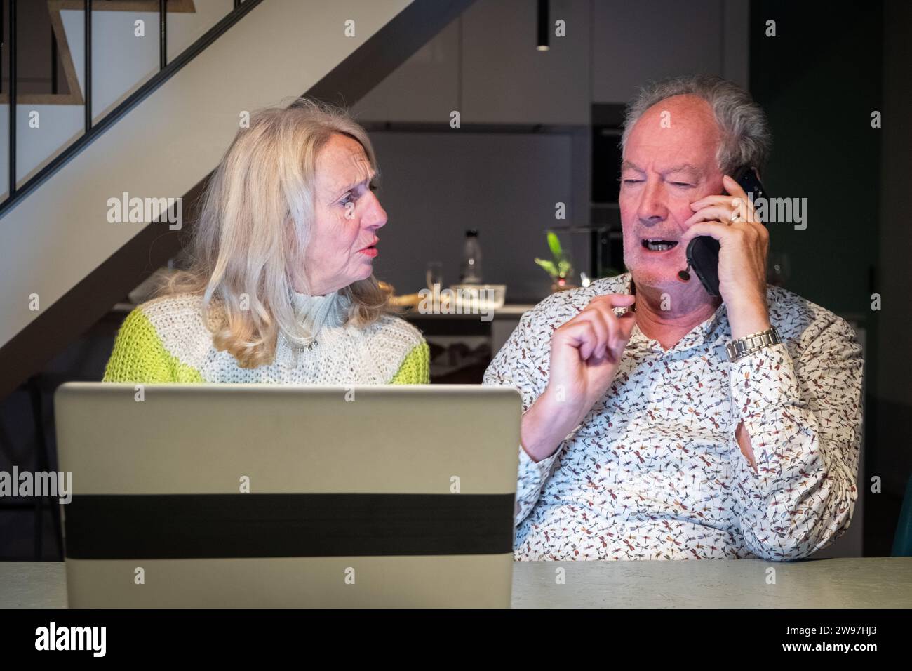 Cette image représente un couple de personnes âgées travaillant ensemble pour résoudre un problème, avec la femme ayant l'air préoccupée ou confuse pendant que l'homme est au téléphone, parlant peut-être avec le support client. L’homme fait des gestes comme s’il expliquait la situation, tandis que la femme semble chercher à se rassurer ou à clarifier. Leur effort de coopération dans le contexte d'une cuisine moderne met en évidence l'intersection des générations plus âgées avec la technologie contemporaine et les systèmes de soutien. Collaboration technique senior, résolution de problèmes en action. Photo de haute qualité Banque D'Images