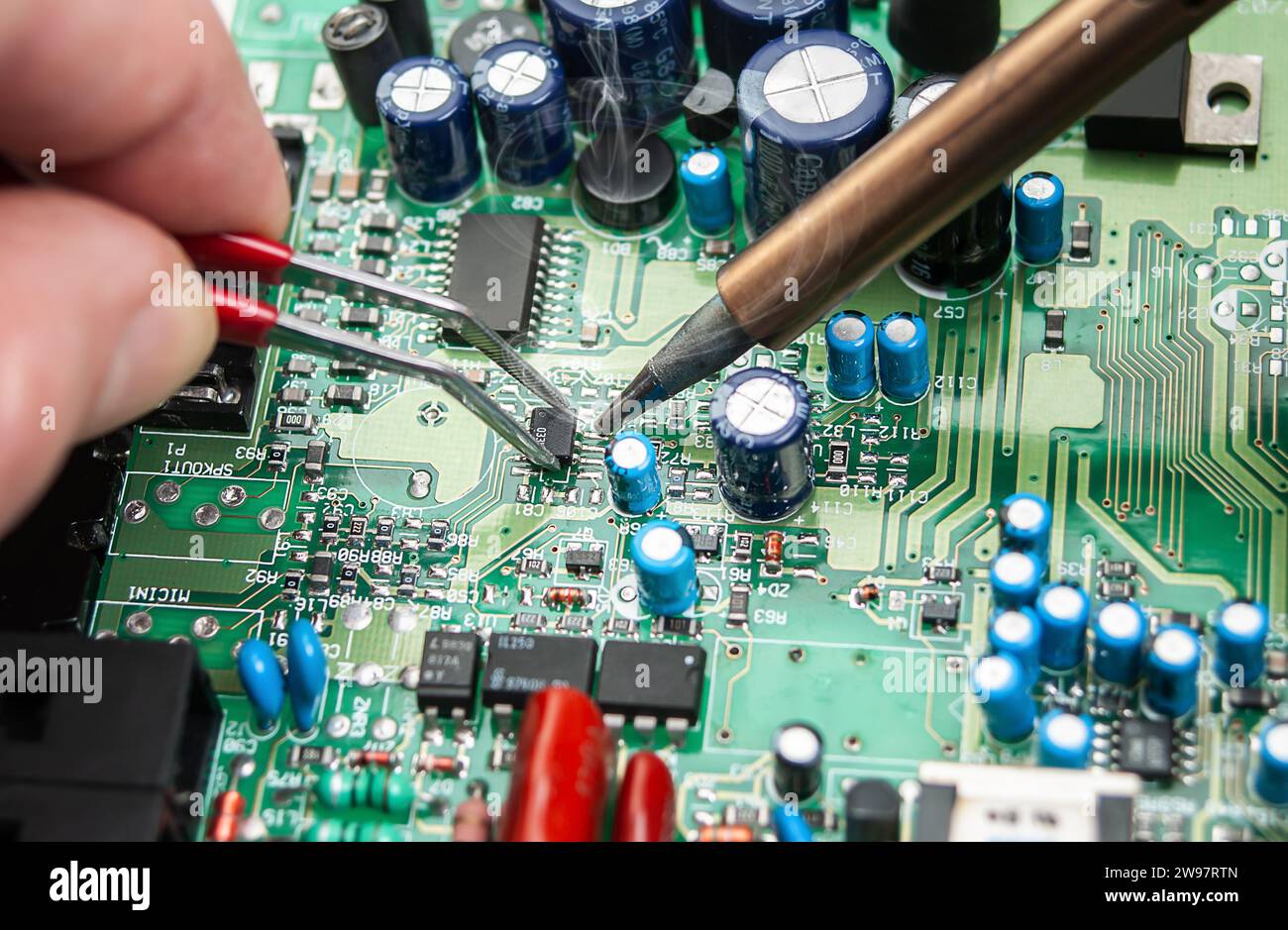 Gros plan des mains d'un technicien dans un atelier. Réparateur soude la carte de circuit imprimé de l'appareil électronique sur la table avec des brucelles. Banque D'Images