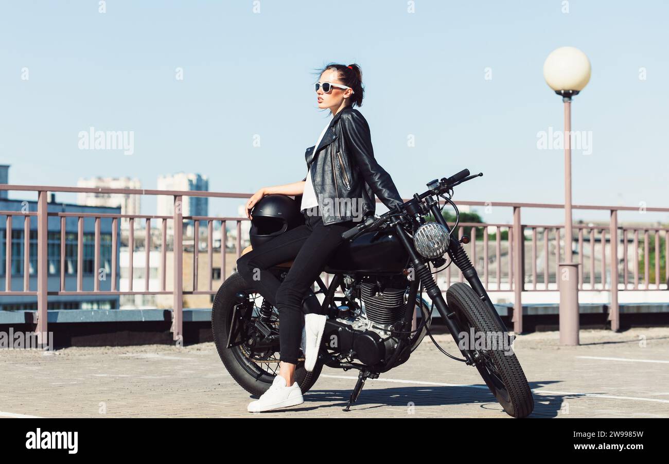 Une jeune femme portant une veste en cuir noir et un pantalon pose avec une moto Banque D'Images