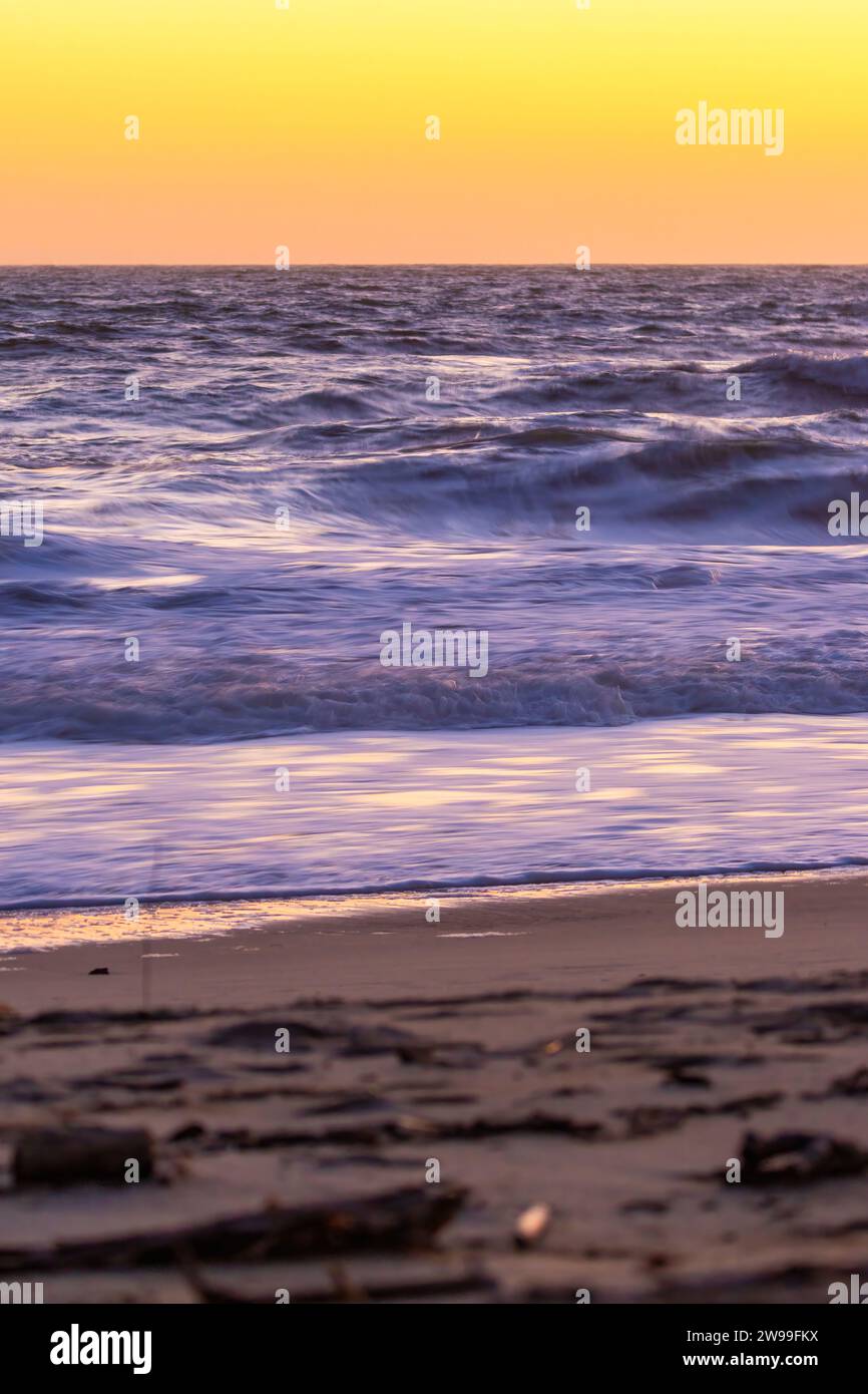 Un coucher de soleil à couper le souffle est capturé sur une plage, avec des vagues qui roulent et un surfeur solitaire marchant dans le sable Banque D'Images