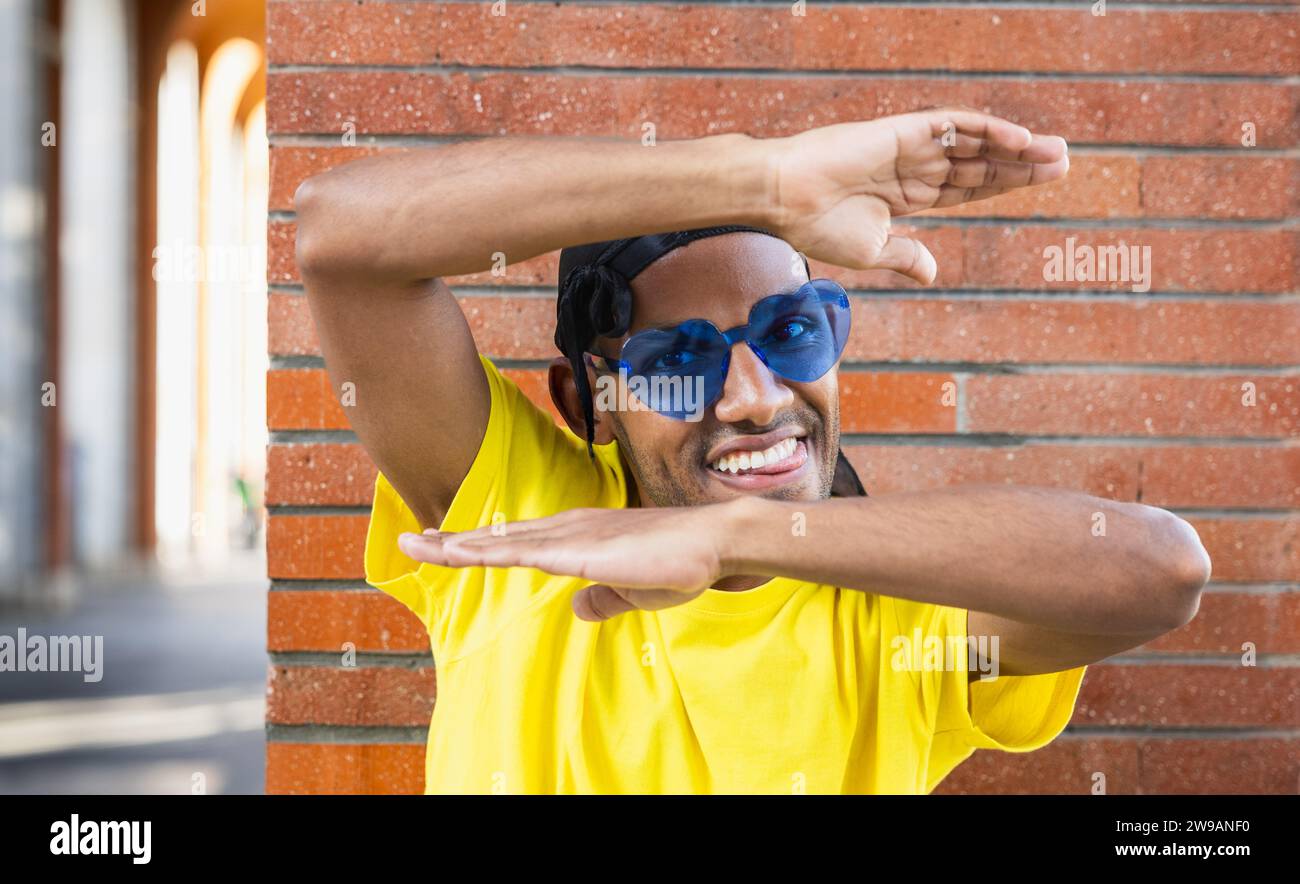 Mec noir joyeux et souriant avec des lunettes colorées encadrant son visage avec ses mains, dans la rue, espace de copie Banque D'Images