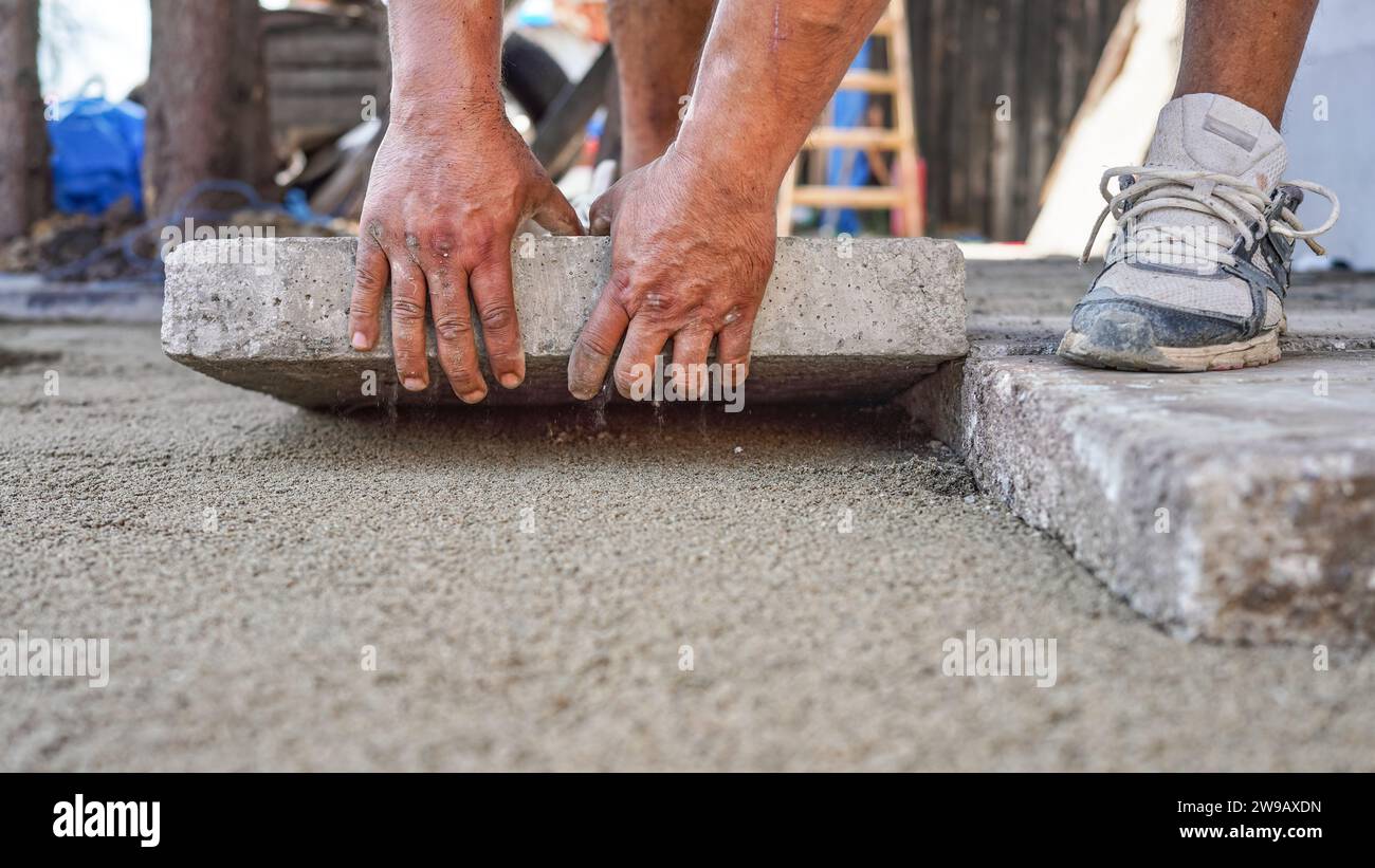 Installation d'une nouvelle chaussée ou d'un nouveau plancher à l'extérieur de grands carreaux de béton, détail de la fermeture sur un travailleur masculin qui pose un bloc de pierre sur une couche de base de sable et de gravier. Banque D'Images