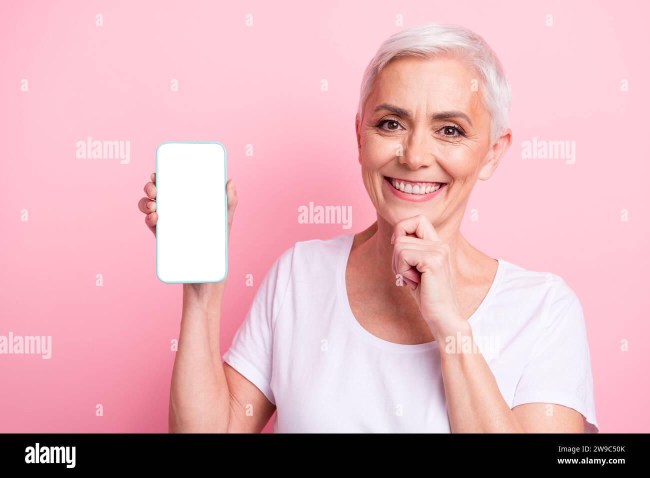 Photo de femme âgée intelligente avec coiffure courte porter blanc t-shirt touch menton montrer écran de smartphone isolé sur fond rose Banque D'Images