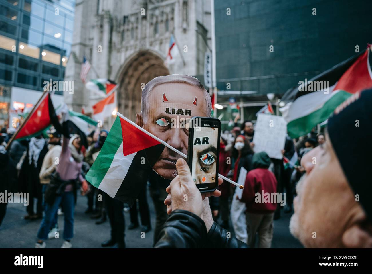 Un manifestant pro-palestinien prend une photo de l'un des manifestants portant un masque représentant le Premier ministre israélien Netanyahu pendant la manifestation. Le jour de Noël, après avoir construit une scène de nativité avec des décombres, du faux sang et des poupées inusées, des centaines de partisans de la Palestine à New York descendent dans la rue pour appeler à un cessez-le-feu immédiat à Gaza. La manifestation est organisée en solidarité avec les églises chrétiennes de Bethléem, qui ont annulé les célébrations de Noël en raison du bombardement continu de Gaza. Banque D'Images