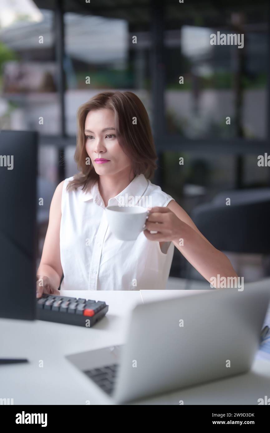 Femmes d'affaires asiatiques boire du café et taper des informations sur le clavier à travailler sur la planification du rapport de marketing sur l'ordinateur tout en analysant investissm Banque D'Images