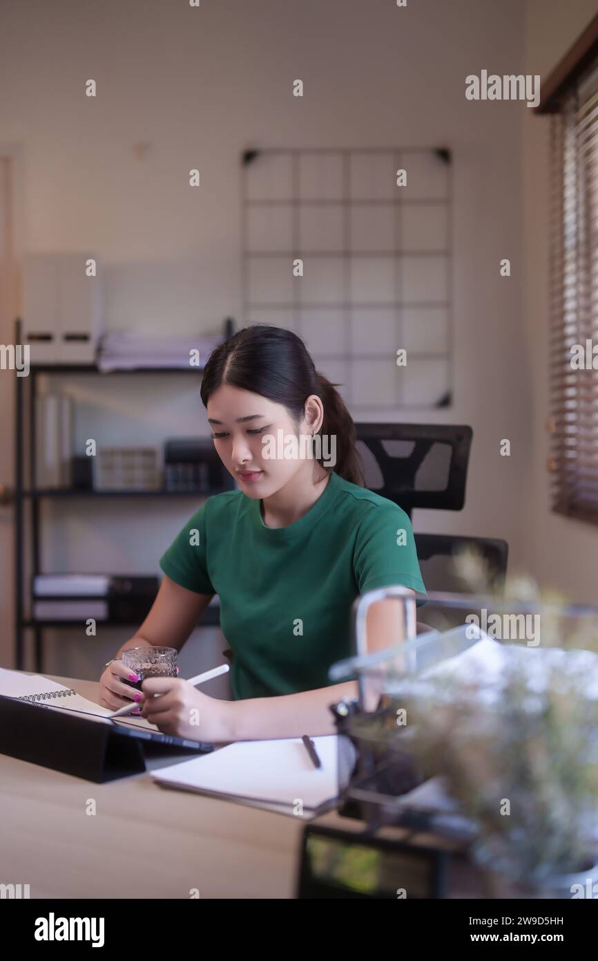 Jeunes étudiantes asiatiques tenant un verre à café et écrivant des notes dans une tablette numérique pour faire leurs devoirs après avoir étudié le webinaire de conférence en ligne an Banque D'Images