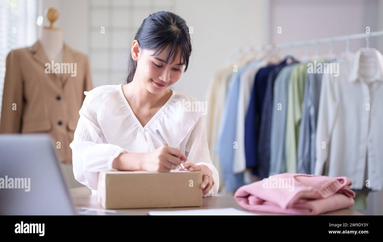 Jeunes femmes marchands asiatiques écrivant les données d'adresse dans la boîte en carton et se préparant à expédier le client de livraison tout en vérifiant l'achat en ligne commande sur l Banque D'Images