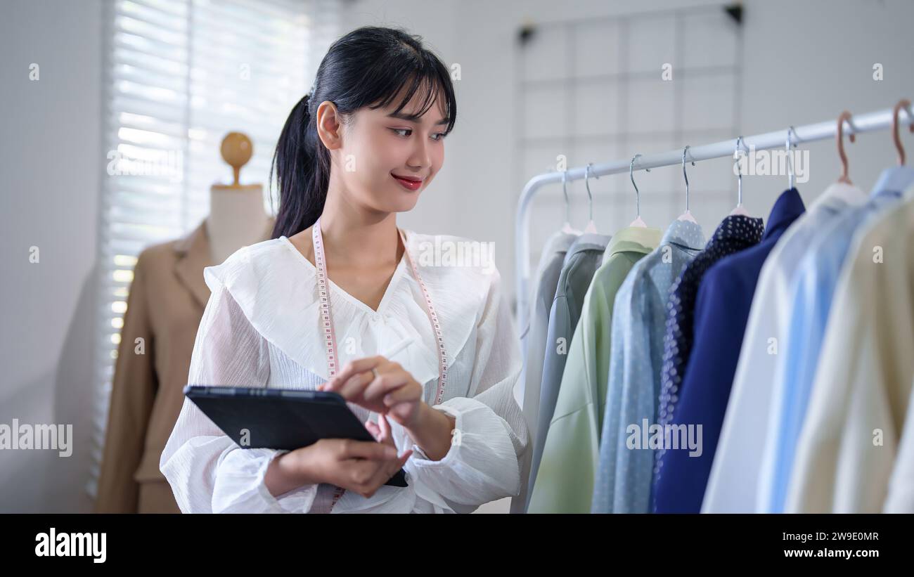Jeunes femmes marchandes asiatiques vérifiant les produits de vêtements stock en magasin et les informations de commande en ligne des clients sur tablette numérique tout en prenant des notes et le travail Banque D'Images