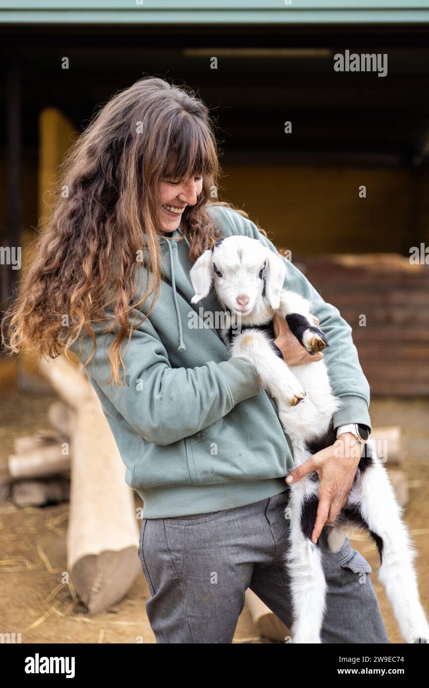 Heureuse jeune femme fermière embrassant un bébé chèvre sur une ferme biologique rurale. Bien-être et soins des animaux dans une grange. Banque D'Images