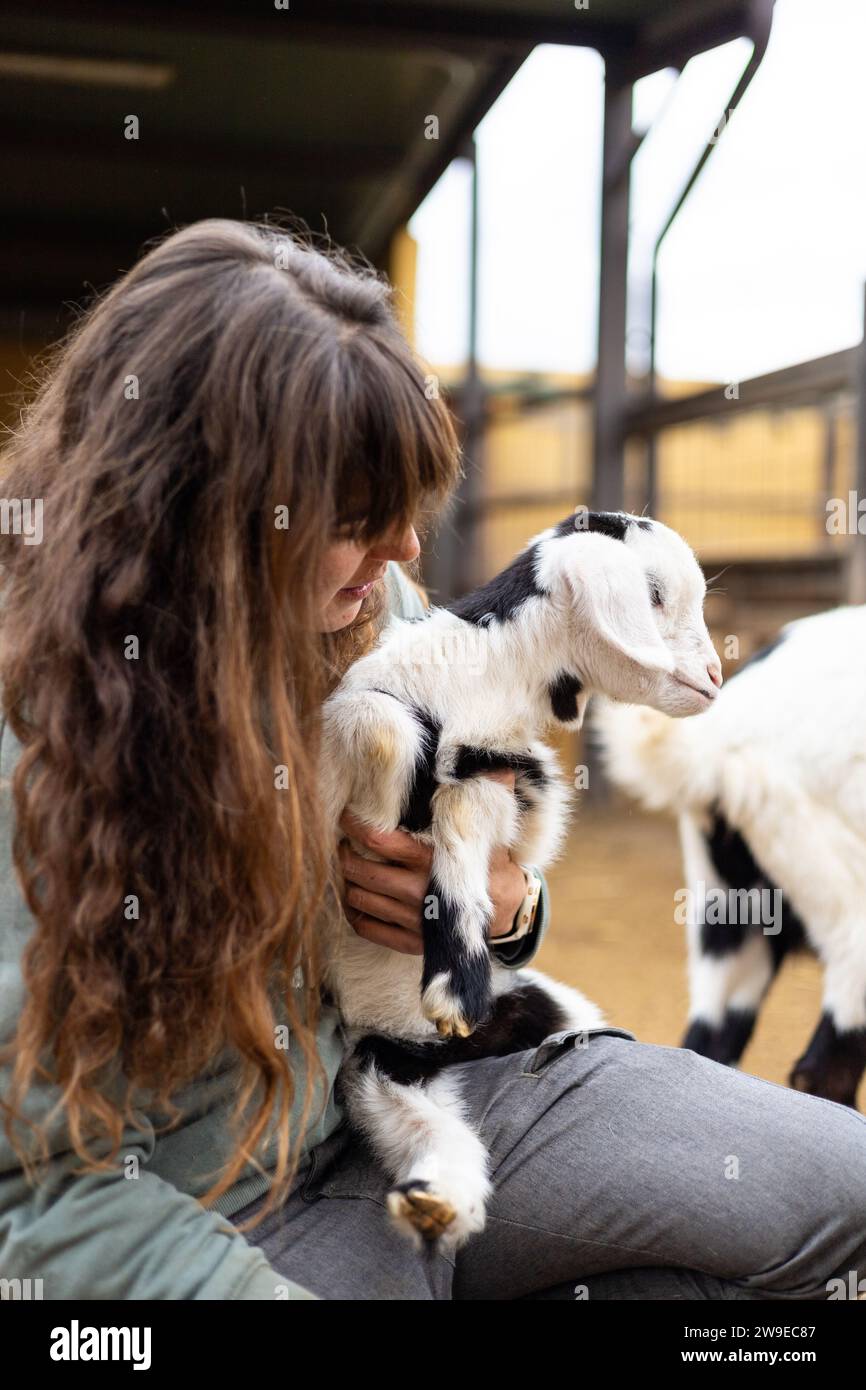 Heureuse jeune femme fermière embrassant un bébé chèvre sur une ferme biologique rurale. Bien-être et soins des animaux dans une grange. Banque D'Images