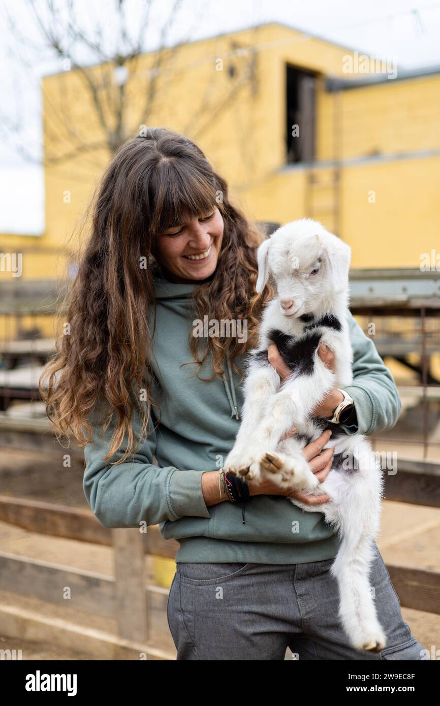 Heureuse jeune femme fermière embrassant un bébé chèvre sur une ferme biologique rurale. Bien-être et soins des animaux dans une grange. Banque D'Images