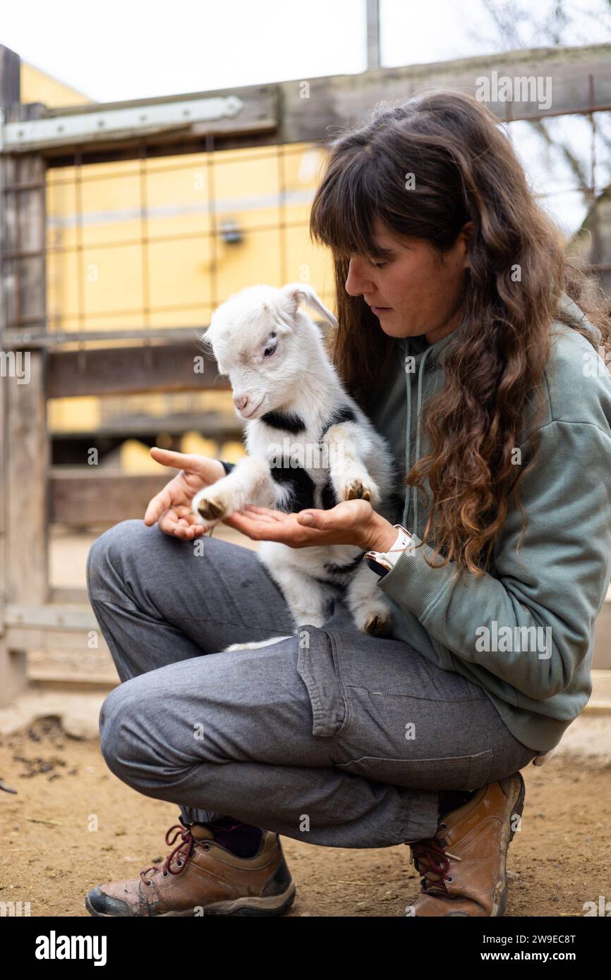 Heureuse jeune femme fermière embrassant un bébé chèvre sur une ferme biologique rurale. Bien-être et soins des animaux dans une grange. Banque D'Images