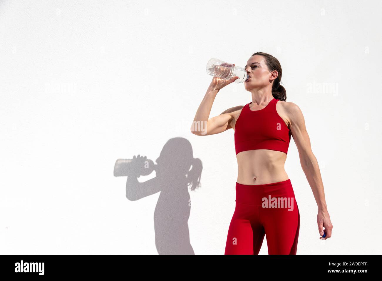 Une femme sportive boit de l'eau après l'exercice et coule à l'extérieur par un mur blanc Banque D'Images