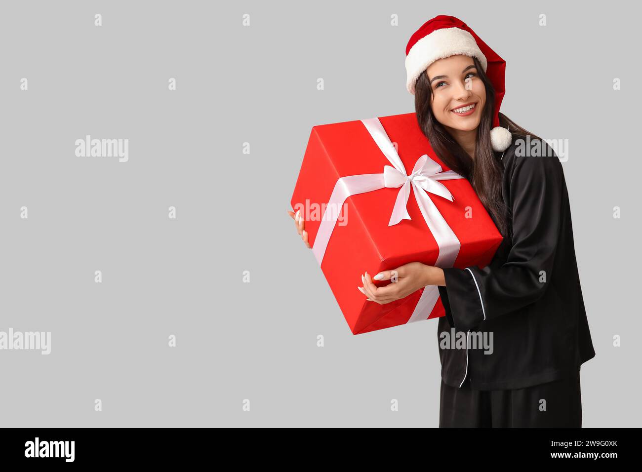 Belle jeune femme en pyjama noir et chapeau de Père Noël avec boîte cadeau de Noël sur fond gris Banque D'Images