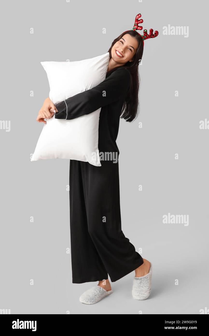 Belle jeune femme en pyjama noir et cornes de renne avec oreiller doux sur fond gris Banque D'Images