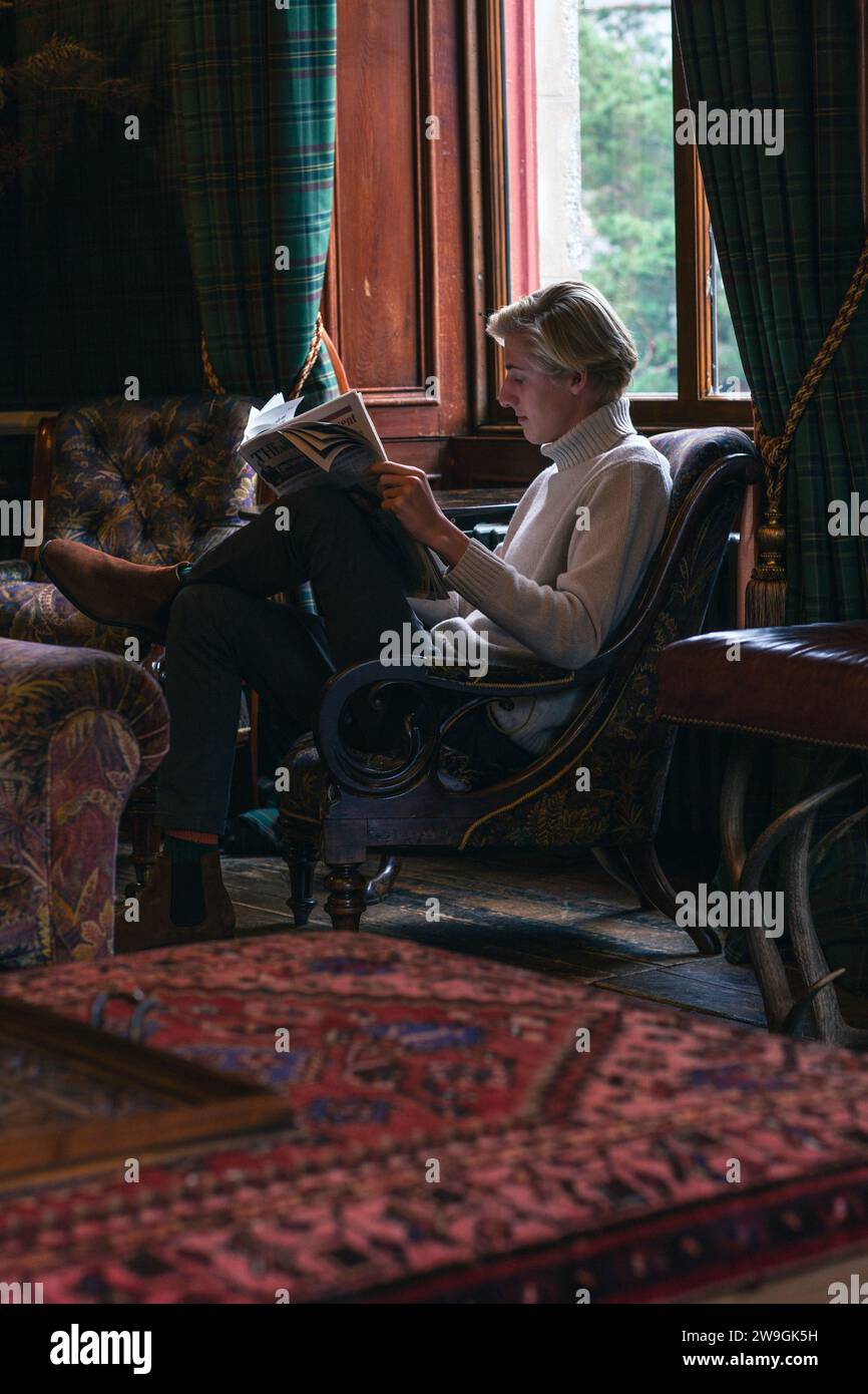 Jeune homme assis dans un fauteuil lisant le journal à l'hôtel The Fife Arms à Braemar , en Écosse . Banque D'Images