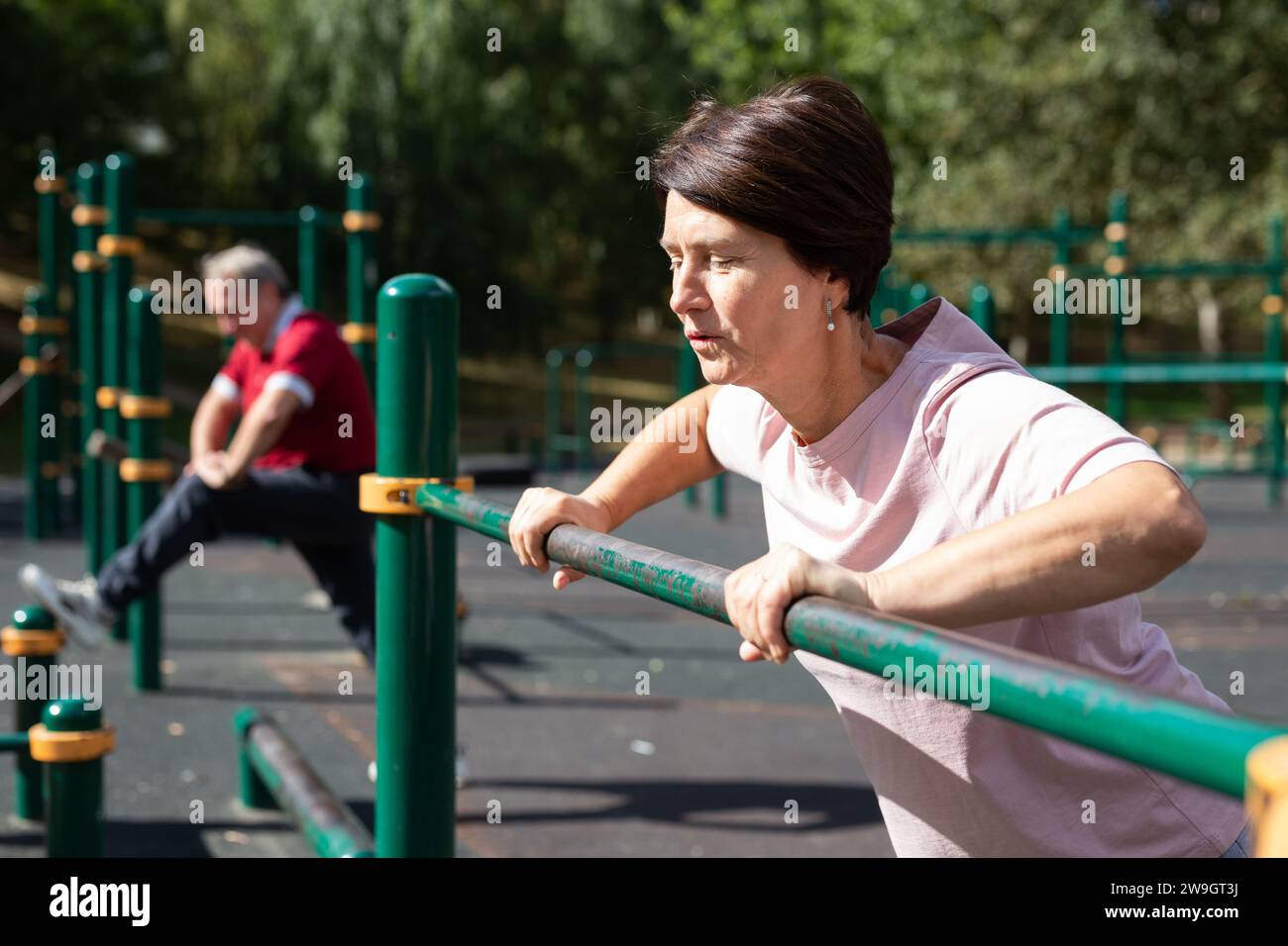 Femme âgée vigoureuse faisant des exercices du matin sur les barres de sport dans le terrain de sport en plein air. Homme faisant du sport en arrière-plan Banque D'Images