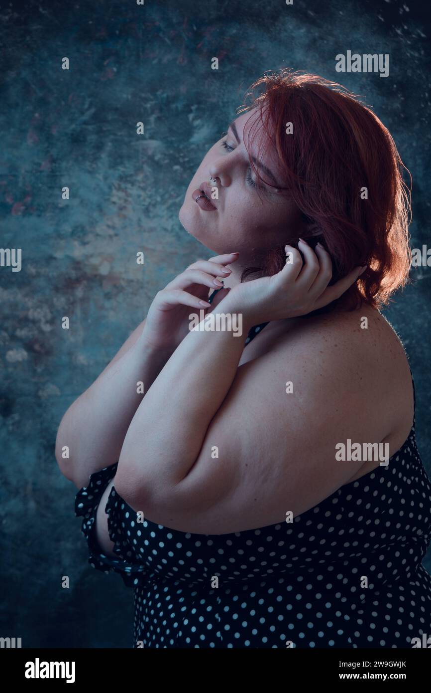 portrait artistique vertical d'une jeune femme latine de taille plus avec la peau blanche, avec les cheveux rouges courts, debout posant avec le fond bleu illuminé Banque D'Images