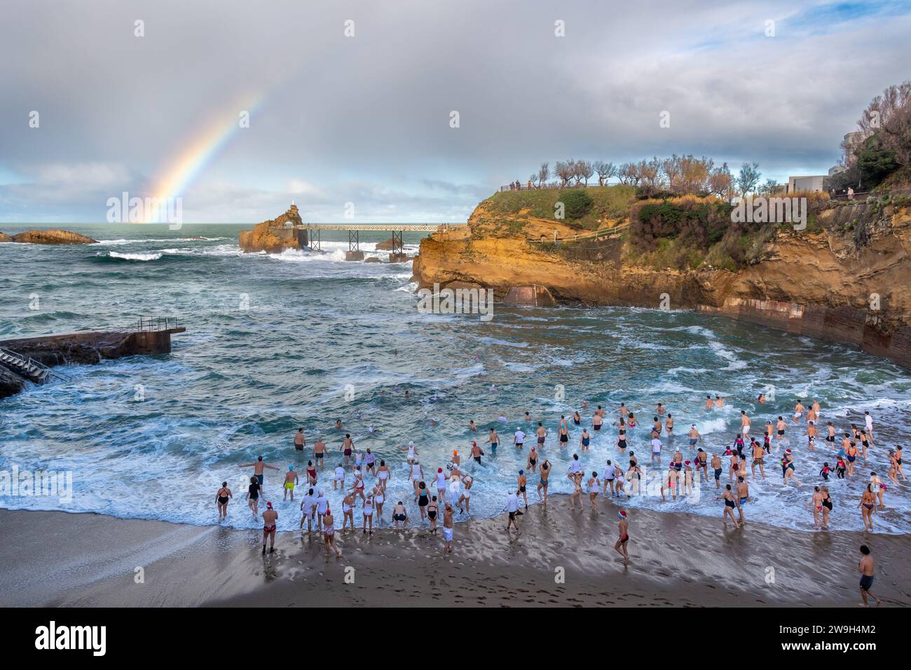 Foule de nageurs entrant dans l'eau pour la traditionnelle baignade de Noël à la plage de Port-Vieux avec un bel arc-en-ciel au-dessus de l'océan Atlantique Banque D'Images