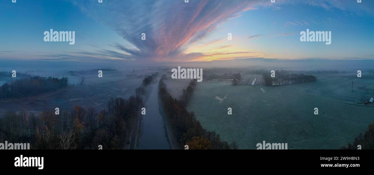 Un panorama grandiose se déploie dans cette photographie aérienne, où la lumière d'un nouveau jour orne doucement un paysage pastoral. Une rivière centrale ou une voie navigable traverse le cadre, menant le regard vers l'horizon où l'aube se lève dans une symphonie de couleurs. Les champs environnants, touchés par la lumière précoce, sont partiellement voilés par une douce brume, ce qui ajoute une qualité onirique au paysage. Le ciel au-dessus est strié de nuages vibrants qui semblent faire écho au chemin de la rivière, créant une symétrie naturelle. Dawn's Embrace sur une étendue de campagne brumeuse. Photo de haute qualité Banque D'Images