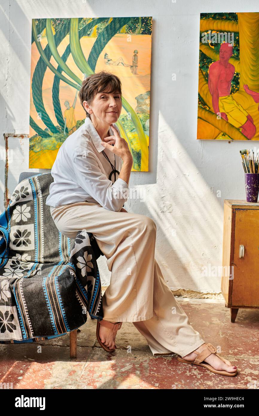 artiste femme d'âge moyen tendance assise sur un fauteuil et souriant à la caméra dans un studio spacieux moderne Banque D'Images