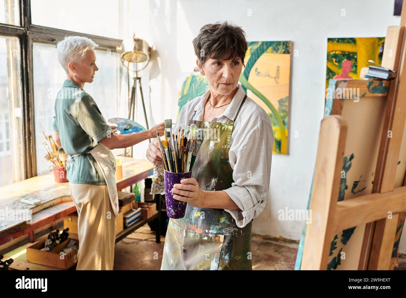 femme mature talentueuse avec des pinceaux regardant le chevalet près de l'amie féminine dans l'atelier d'art. Banque D'Images