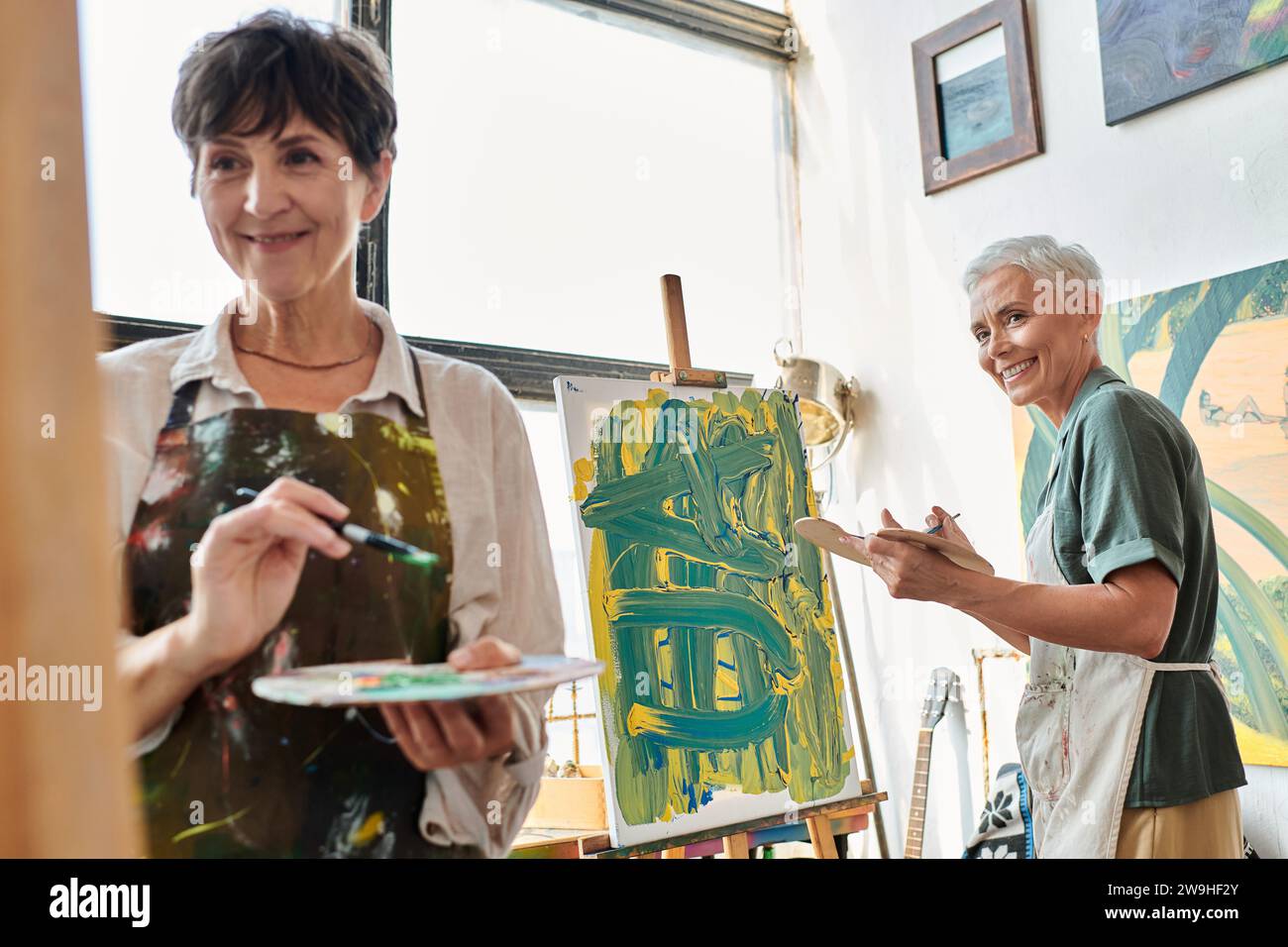 femme mature heureuse avec palette regardant l'atelier d'art de peinture d'amie féminine, passe-temps créatif Banque D'Images