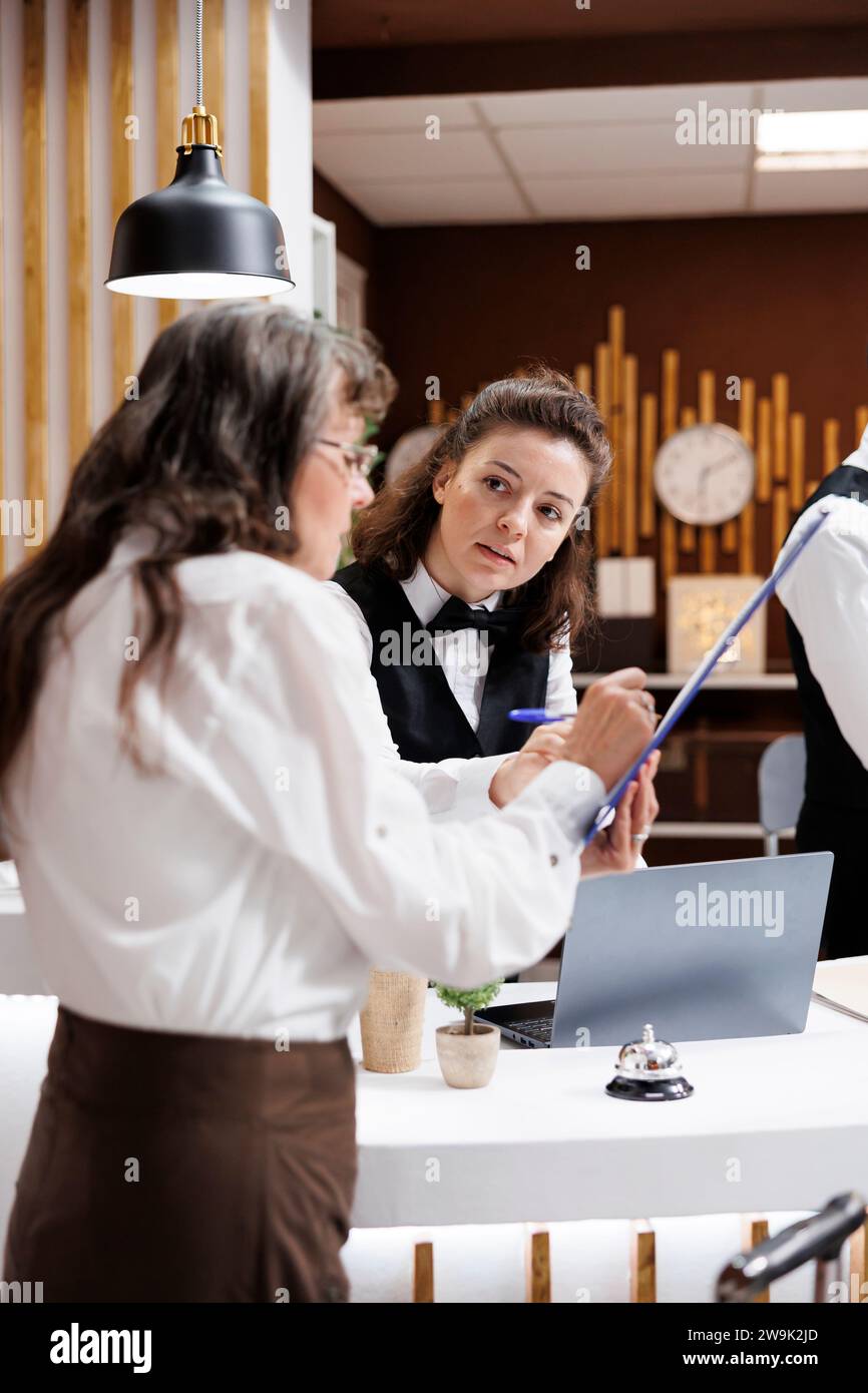 Un concierge professionnel assiste une femme âgée à la retraite à la réception de l'hôtel, assurant un processus d'enregistrement en douceur. La réceptionniste offre un service amical aux voyageurs âgés qui signent des formulaires de réservation. Banque D'Images