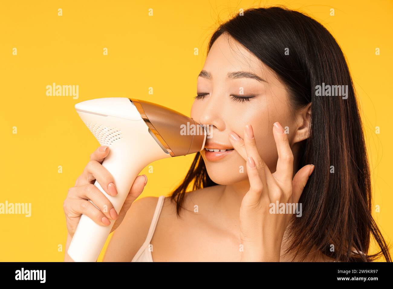 Jolie femme asiatique utilisant photoépilateur sur son visage sur fond jaune Banque D'Images