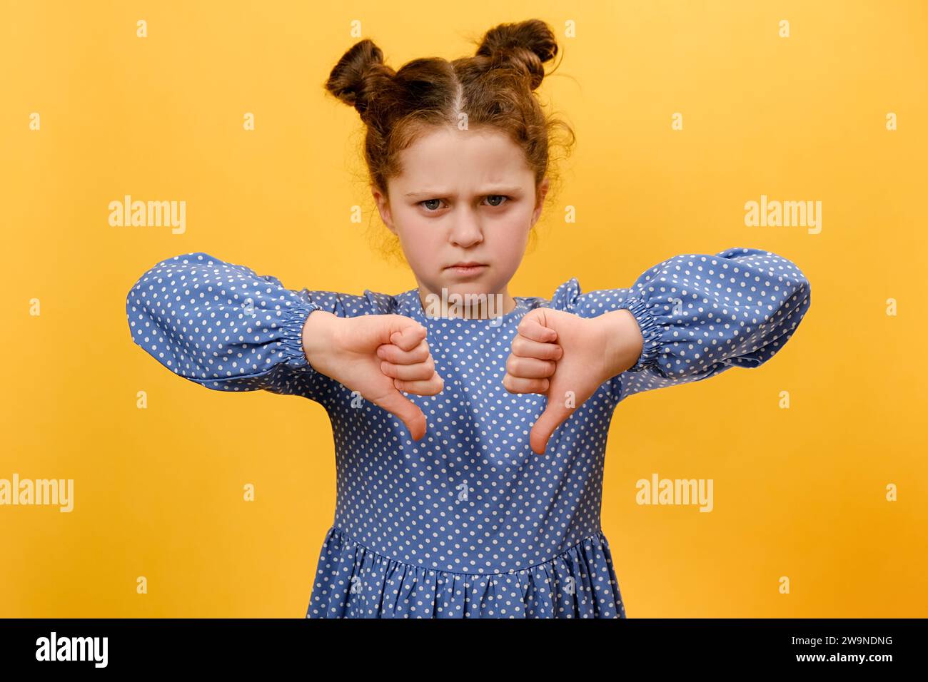 Portrait d'une fille préadolescente coquine faisant des gestes pouces vers le bas avec les deux mains et grimacant, posant isolé sur le mur de fond de couleur jaune dans le studio. Emoti Banque D'Images