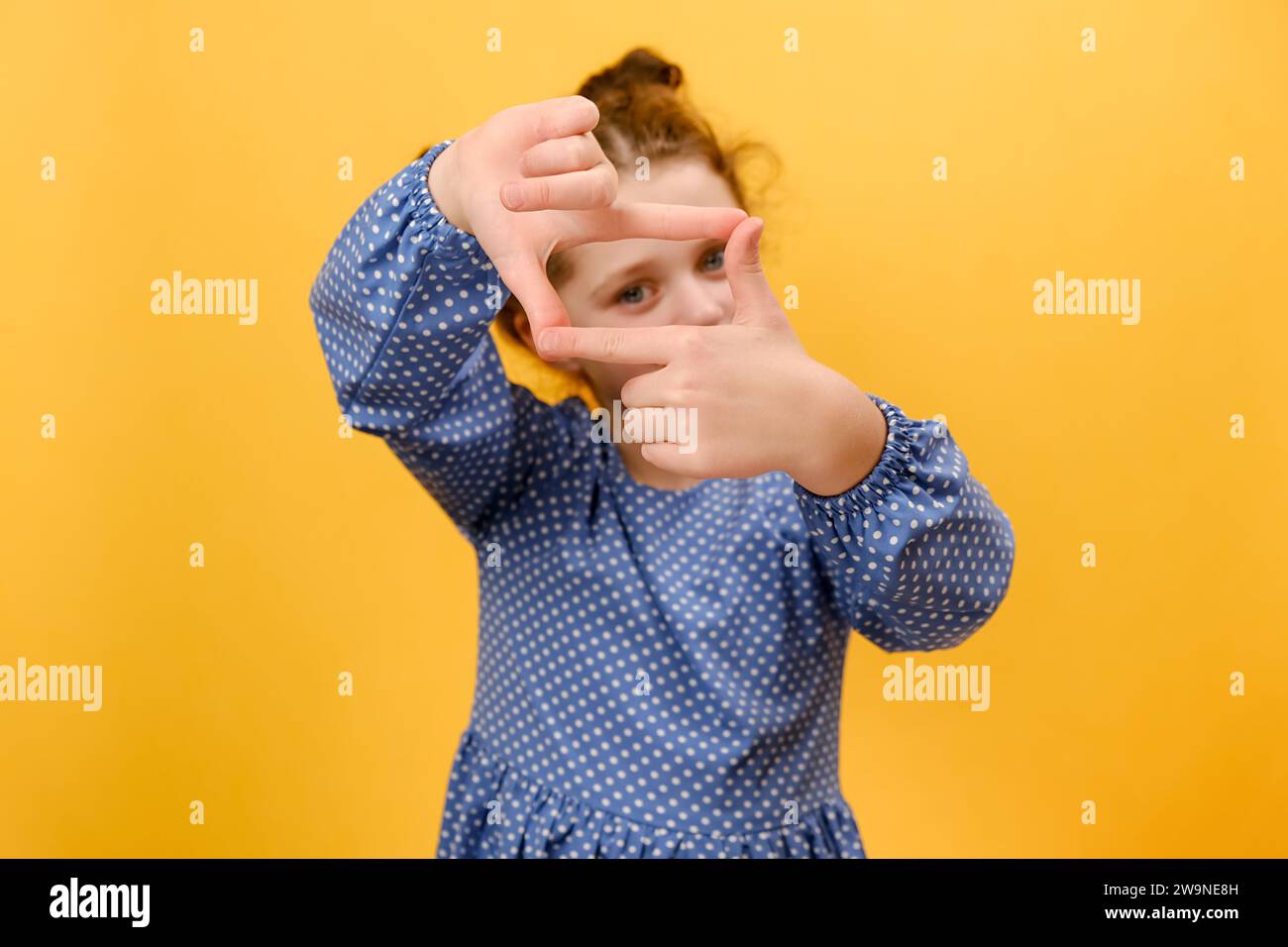 Portrait de mignonne petite fille faisant cadre avec les doigts, regardant la caméra, faisant semblant de faire de la photographie, posant isolé sur fond de couleur jaune Banque D'Images