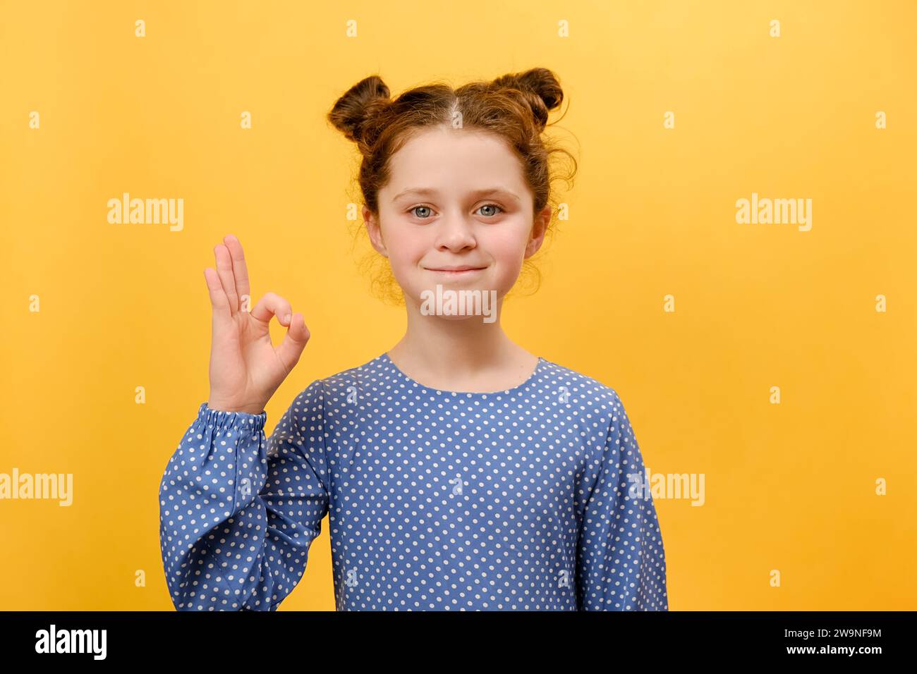 Portrait de la petite fille heureuse enfant montrant le geste OK et souriant regardant la caméra, posant isolé sur le mur de fond de couleur jaune Uni dans le studio wi Banque D'Images