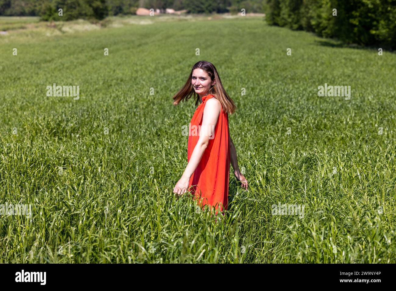 Capturée dans une vaste étendue de champs verdoyants sous un ciel bleu clair, une femme dans une robe orange sans manches frappante se démarque avec son sourire radieux. Elle marche dans le champ avec un toucher doux, sa main paissant les pointes de l'herbe haute. La lumière naturelle de la journée ajoute une lueur chaude à son teint et met en valeur ses cheveux longs et fluides. Son comportement détendu et l'isolement du terrain transmettent un sentiment de liberté et de connexion avec la nature. Cette image dépeint une image de joie estivale et les plaisirs simples d'être à l'extérieur. Femme dans la robe orange vibrante appréciant le Countr Banque D'Images