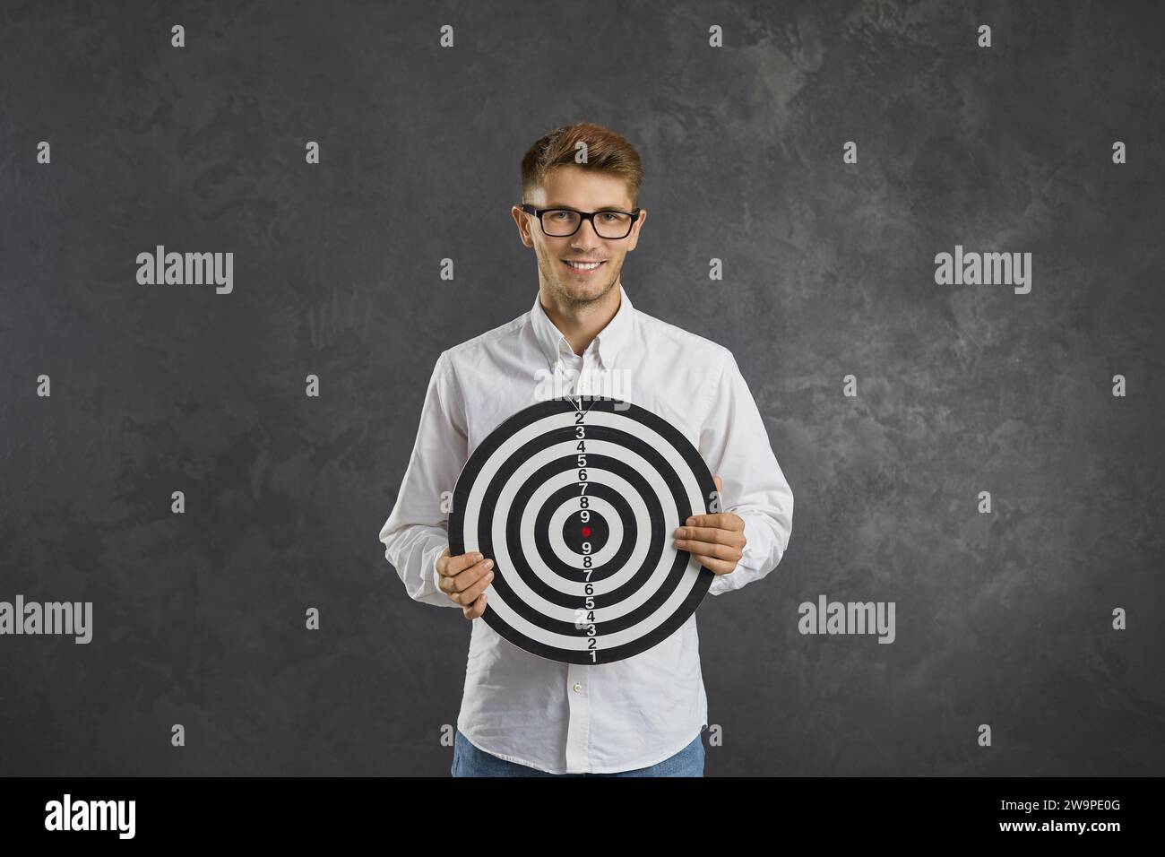 Portrait de jeune homme intelligent et positif tenant le tableau de fléchettes debout sur fond gris. Banque D'Images