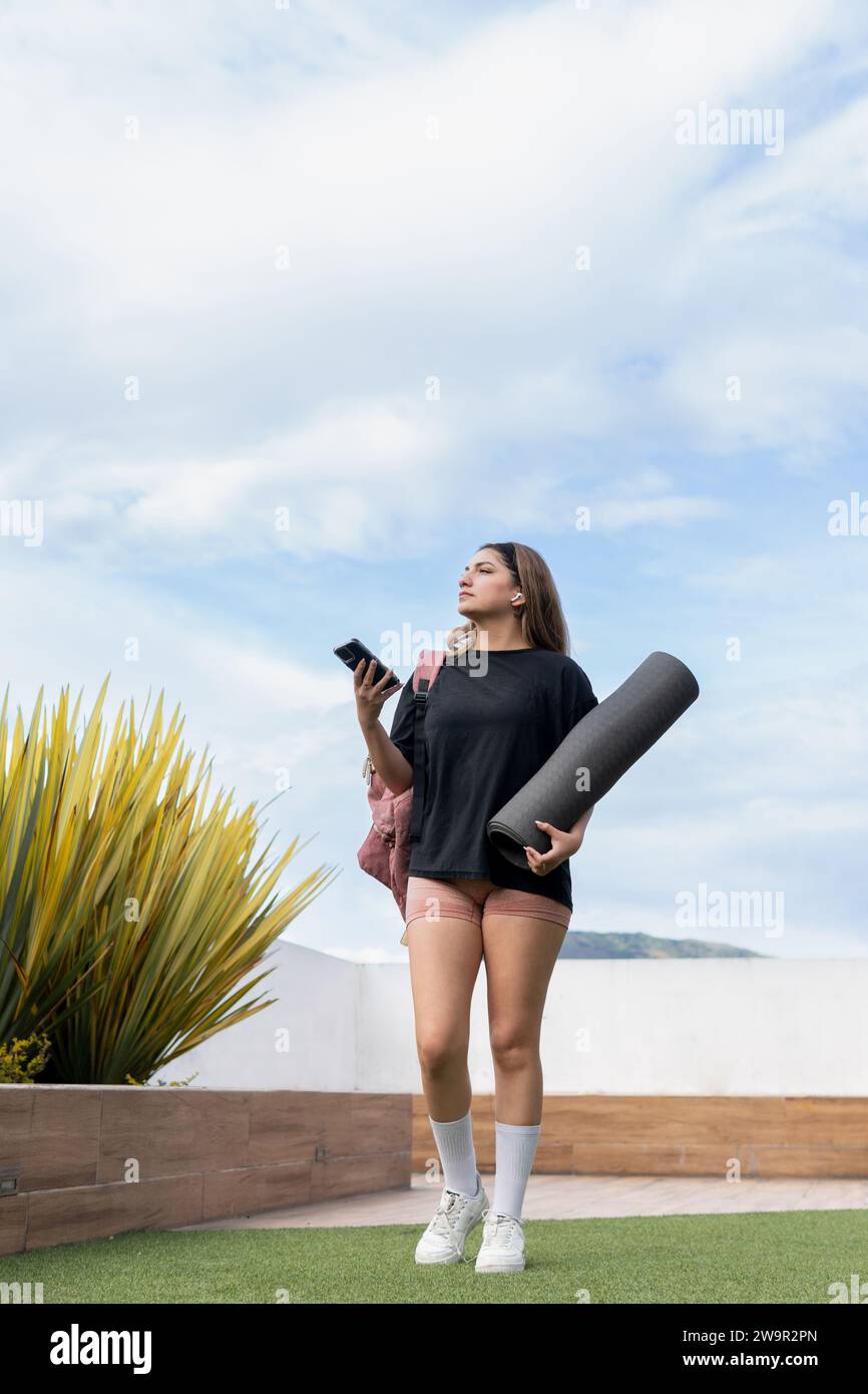Jeune femme Latina portant un short et un t-shirt noir marchant avec un mat et son téléphone portable vers la salle de gym Banque D'Images