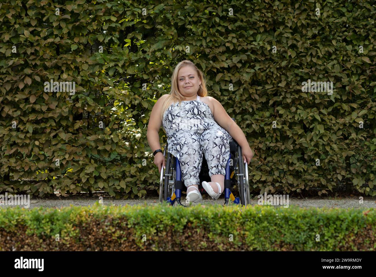Journée internationale des fauteuils roulants , le 1 mars souriant. Jolie jeune femme avec stature courte assise dans fauteuil roulant en plein air. Mur vert sur fond Banque D'Images