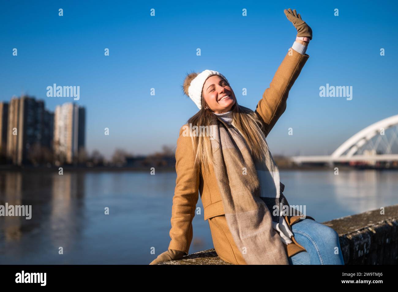 Belle femme en vêtements chauds aime se reposer au bord de la rivière par une journée ensoleillée d'hiver. Banque D'Images