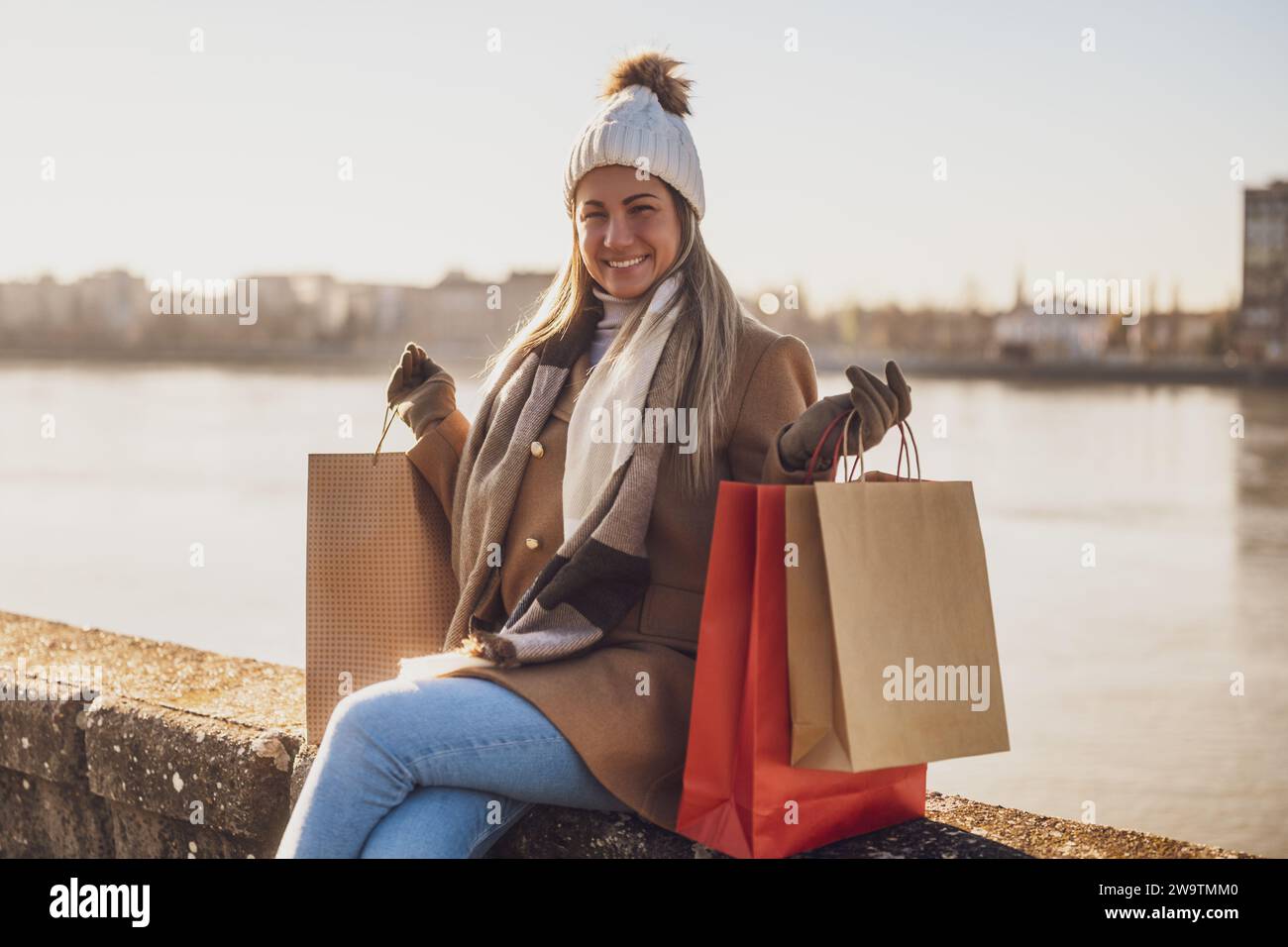 Femme heureuse dans des vêtements chauds avec des sacs à provisions aime assis au bord de la rivière sur la journée ensoleillée d'hiver. Banque D'Images