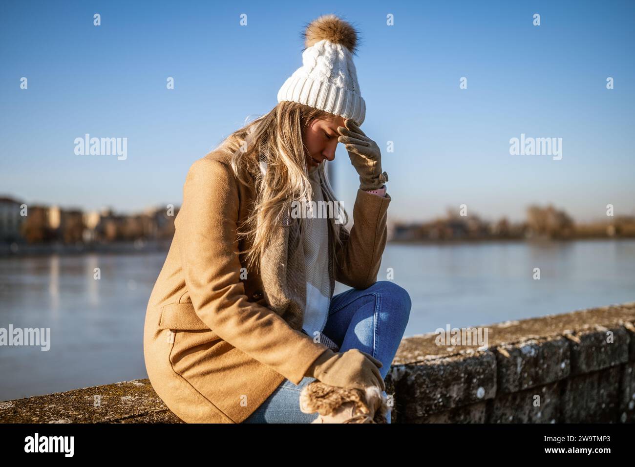 Femme inquiète dans des vêtements chauds ayant mal à la tête et assis au bord de la rivière sur une journée ensoleillée d'hiver. Banque D'Images