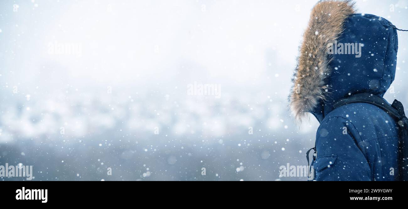 Vue arrière du voyageur dans une veste bleue avec une capuche en fourrure et un sac à dos sur le fond d'un paysage hivernal pendant les chutes de neige Banque D'Images