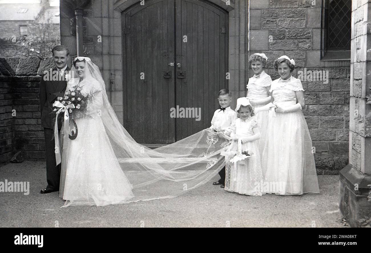 Années 1950, historique, mariage, en dehors de l'église, un petit pageboy et une jeune fille de fleur tiennent le train de la mariée alors qu'elle se tient avec son nouveau mari, Angleterre, Royaume-Uni, avec ses deux demoiselles d'honneur en présence. Banque D'Images