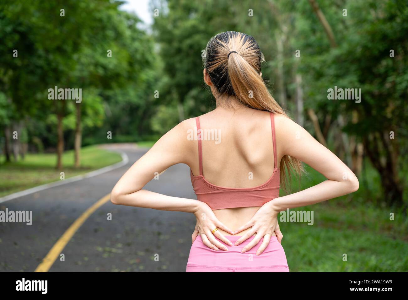 Vue arrière d'une femme aux cheveux longs avec mal de dos mettant ses mains sur son mal de dos bas tout en se tenant sur une piste de course d'un parc local Banque D'Images
