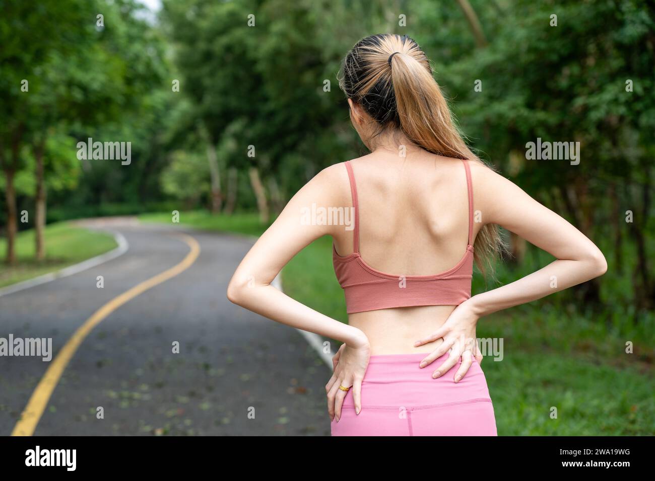 Vue arrière d'une femme aux cheveux longs avec mal de dos mettant ses mains sur son mal de dos bas tout en se tenant sur une piste de course d'un parc local Banque D'Images