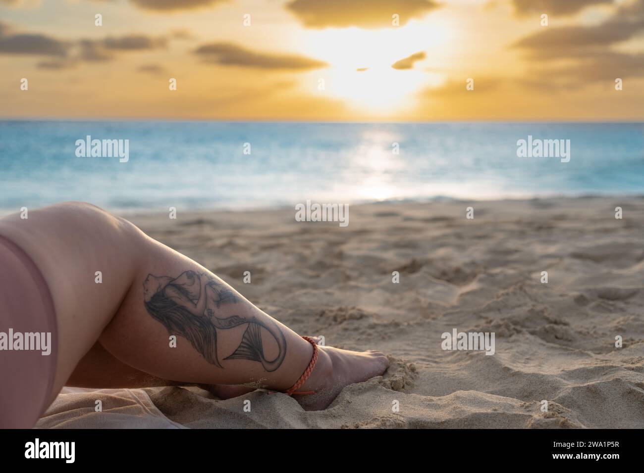 Tatouage de sirène sur une jambe de femme, alors que le soleil se couche sur la couleur bleue des lueurs de l'océan. Banque D'Images